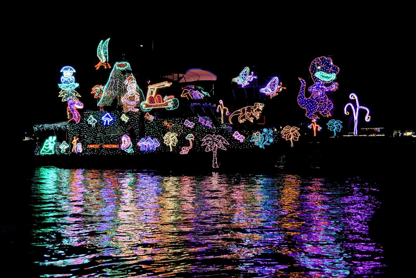 Boat covered in light-up dinosaurs, palm trees and a volcano, reflected in dark water